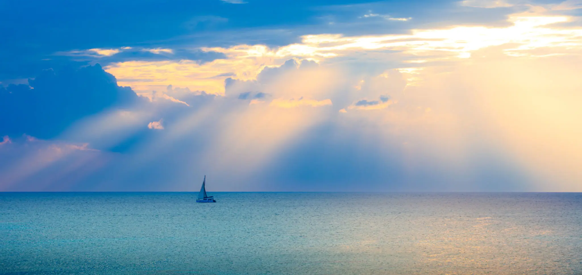 A sailboat on calm sea under dramatic cloud rays.