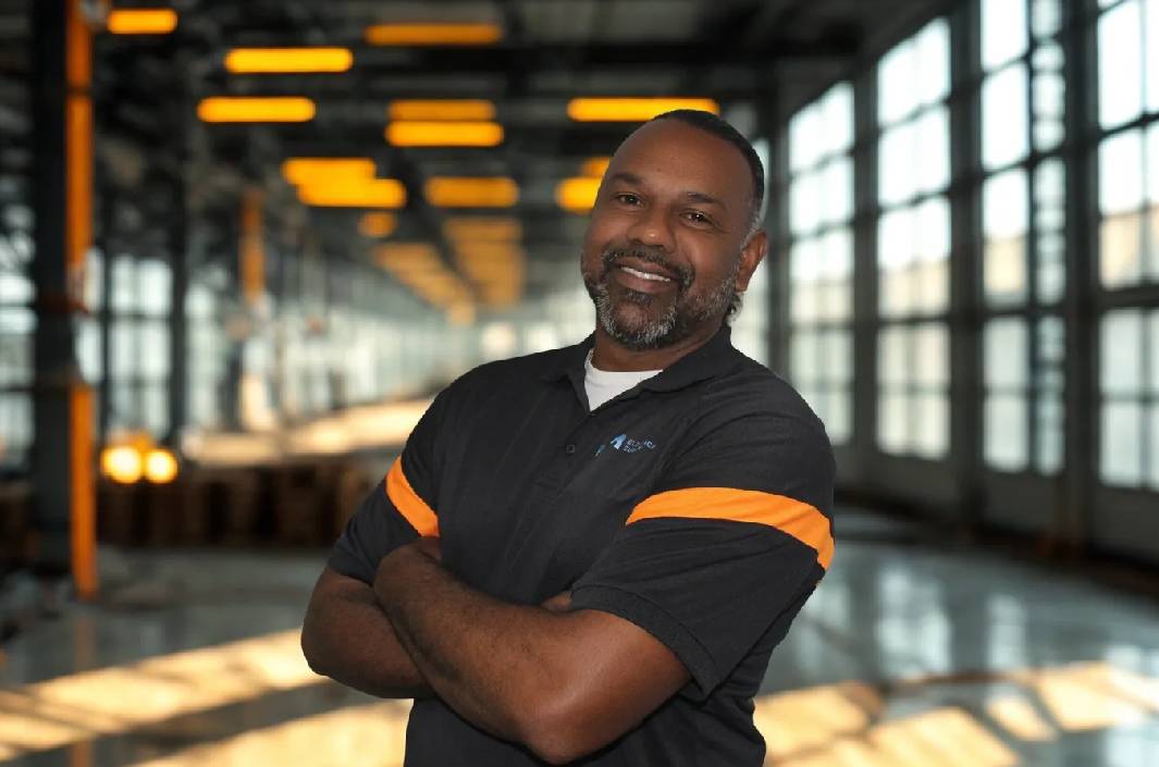 Confident man in a black shirt with orange stripes, arms crossed, smiling in a modern, industrial setting.